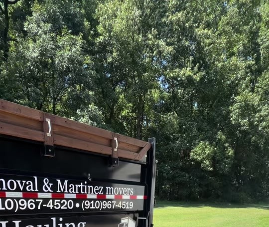 Dump trailer lettered for Junk Removal and Martinez Movers parked beside a wooded lawn